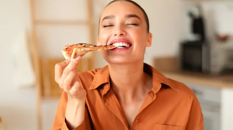 Frau isst Pizza und lächelt Frau in Küche beißt lachend in ein Stück Pizza.